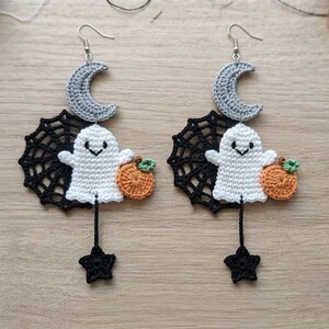 Puede incluir: Un par de pendientes de ganchillo con una luna creciente gris en la parte superior, un fantasma blanco con cara sonriente, una calabaza naranja, un fondo de telaraña negra y una pequeña estrella negra colgando de un gancho plateado.