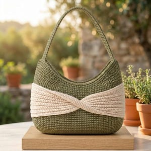 May include: An olive green crochet handbag featuring a single shoulder strap and a wide, cream-coloured horizontal band across the centre that is twisted to create a knot-like detail. The bag has a structured, rounded shape with a textured surface.