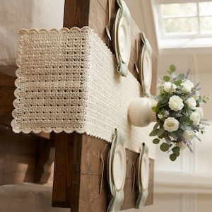 May include: A cream-colored rectangular crochet table runner with a scalloped edge, laid across a dark wooden table. The table is set with four white plates, each topped with a folded light green napkin, and a white vase filled with white roses.