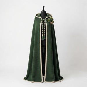May include: A floor-length dark green hooded cloak featuring a cream-colored trim along the edges. The garment includes a braided tie closure with tassels and a decorative cluster of fabric oak leaves and small brown acorns attached at the shoulder.