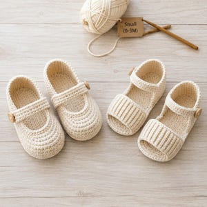 Puede incluir: Dos pares de zapatos de ganchillo color crema con botones de madera. Un par tiene un diseño cerrado, mientras que el otro es un estilo de sandalia con punta abierta. Un ovillo de lana y una etiqueta que dice Small (0-3M) están arriba.