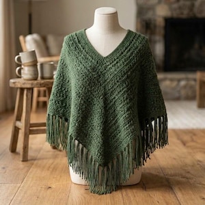 May include: A forest green crochet poncho featuring a V-neckline and a textured stitch pattern. The garment has a pointed hemline finished with long, matching green fringe trim that hangs from the bottom edges of the knitted fabric.