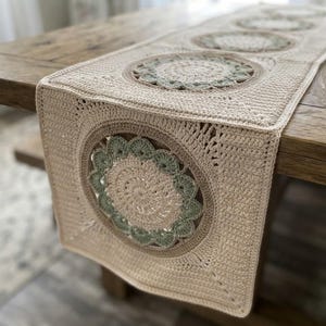 May include: A rectangular crochet table runner featuring a series of repeating square motifs. Each square contains a central circular design in cream and sage green yarn, framed by a solid cream border with a textured, hand-stitched appearance.