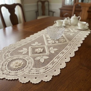 Op de afbeelding: Een gebroken witte, ovale tafel loper van haakwerk met ingewikkelde bloemmotieven, geometrische vierkante patronen en een geschulpte rand. De loper is gemaakt van fijn garen en toont gedetailleerd handwerk en een delicaat opengewerkt kantontwerp.