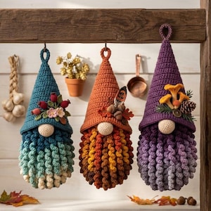 Pode incluir: Três gnomos de crochê pendurados com chapéus pontiagudos e barbas encaracoladas. O gnomo da esquerda tem um chapéu azul-petróleo com bagas e flores. O central tem um chapéu laranja com bolotas e folhas. O da direita tem um chapéu roxo.
