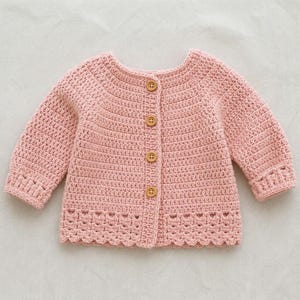 May include: A light pink long-sleeve cardigan made of crocheted yarn. The garment features a round neckline, four small brown wooden buttons down the centre front, and a decorative scalloped crochet pattern along the bottom hem and cuffs.