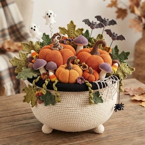 May include: A crochet cauldron filled with orange pumpkins, purple mushrooms, green leaves, and candy corn. The arrangement features white ghost figures, grey bats on wires, and a small black spider hanging from a white crocheted spider web.