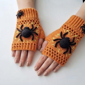 May include: A pair of orange fingerless gloves featuring a crocheted texture with a ribbed cuff. Each glove is decorated with a large, three-dimensional black spider appliqué and a smaller black spider, accented by yellow embroidered starbursts.