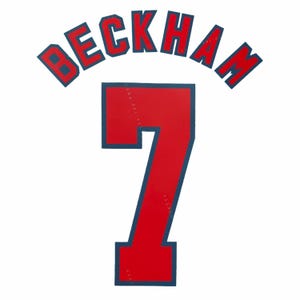 May include: A red and dark blue iron-on transfer featuring the name BECKHAM in an arched block font above the large number 7. The design uses bold red lettering and numerals with a dark blue outline, intended for application onto a sports jersey.