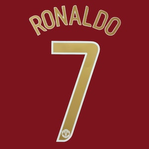 May include: A dark red sports jersey featuring the name RONALDO in gold lettering arched above a large gold number 7. The number 7 has a white outline and includes a small Manchester United team crest logo at the base of the digit.