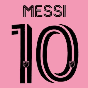 May include: A pink fabric background featuring the black text MESSI above the large black number 10. The number 10 includes a stylized double-line design and features a small MLS logo emblem at the base of both the number one and the zero.