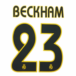 May include: A black and yellow sports jersey name and number set featuring the name BECKHAM in a bold font above the large number 23. Each digit of the number 23 contains a small yellow Real Madrid club crest logo at the bottom base.
