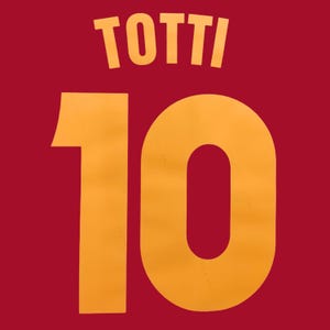 May include: A dark red sports jersey fabric featuring the name TOTTI printed in a bold, yellow sans-serif font above the large number 10, also rendered in a matching yellow colour with a clean, block-style typeface design.