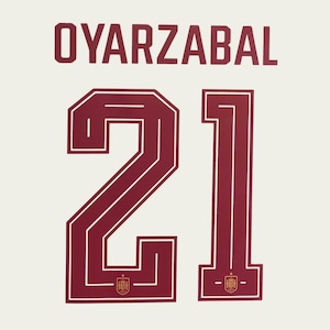 Puede incluir: La parte trasera de una camiseta deportiva blanca con el nombre OYARZABAL en letras mayúsculas de color rojo oscuro sobre el número 21. El número 21 está en una fuente grande de color rojo oscuro con bordes blancos y un escudo español dorado.