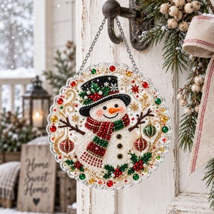 Puede incluir: Un adorno colgante circular que presenta un muñeco de nieve con un sombrero de copa negro con acebo y una bufanda a rayas rojas y verdes. El diseño está adornado con cuentas y cristales facetados de colores, incluyendo adornos rojos y verdes.