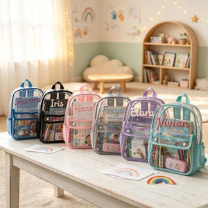 May include: A row of six clear plastic backpacks with coloured trim in blue, black, pink, grey, purple, and teal. Each backpack features a personalised name embroidered on the front pocket: Aurora, Iris, Hazel, Freya, Elara, and Vivian.