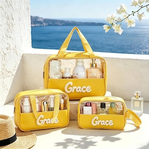 Puede incluir: Tres neceseres rectangulares amarillos con paneles frontales de pl&aacute;stico transparente y asas superiores. Cada bolsa presenta el nombre Grace en letras cursivas blancas texturizadas en el frente, rellenas con varios frascos y recipientes.