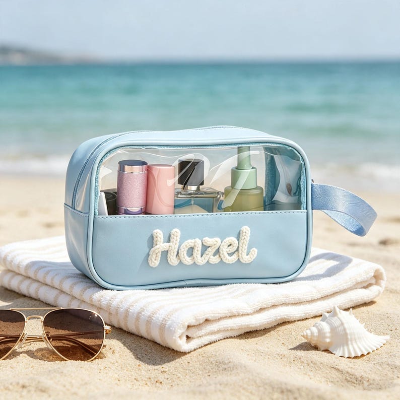 Puede incluir: Un neceser rectangular de color azul claro con un panel frontal transparente y un asa lateral. El nombre Hazel est&aacute; bordado en letra blanca en la parte delantera. El neceser contiene varios frascos de cosm&eacute;ticos sobre una toalla blanca.