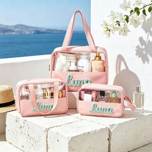 Puede incluir: Tres neceseres de color rosa claro con paneles de pl&aacute;stico transparente y el texto Luna bordado en verde menta. Cada bolsa cuenta con asas resistentes y contiene varios frascos de cuidado personal y cosm&eacute;ticos para organizar.