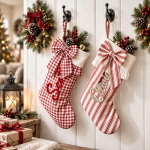 Pode incluir: Duas meias de Natal penduradas em ganchos de parede pretos. A meia esquerda apresenta um padrão xadrez vichy vermelho e branco com um laço a condizer e texto vermelho. A direita tem riscas verticais vermelhas e brancas, um laço e texto prateado.