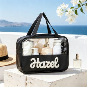 Puede incluir: Un neceser rectangular negro con un panel frontal transparente y asas de tela negra. La parte delantera presenta el nombre Hazel en letras cursivas blancas texturizadas. El bolso contiene varios frascos blancos y beige y una esponja.