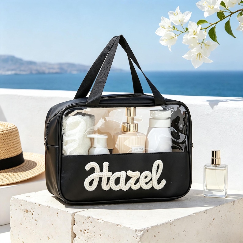 Puede incluir: Un neceser rectangular negro con un panel frontal transparente y asas de tela negra. La parte delantera presenta el nombre Hazel en letras cursivas blancas texturizadas. El bolso contiene varios frascos blancos y beige y una esponja.