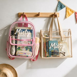 Op de afbeelding: Een doorzichtige plastic rugzak met roze randen en een doorzichtige plastic draagtas met beige randen, beide voorzien van de naam Misty in witte reliëfletters. De tassen bevatten boeken, een etui, pennen, een paraplu en een bril.