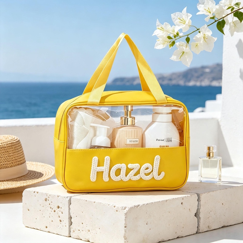 Puede incluir: Un neceser de color amarillo brillante con un panel frontal transparente y dos asas superiores. El nombre Hazel est&aacute; bordado en letra blanca en la parte inferior frontal. Dentro hay tres botellas de productos de cuidado personal.
