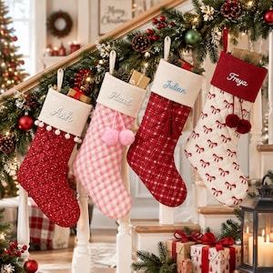 Puede incluir: Cuatro calcetines navideños acolchados colgados de una barandilla de escalera. Los calcetines tienen puños blancos bordados con los nombres Ash, Hazel, Julian y Freya. Los diseños incluyen patrones rojos, cuadros rosas y lazos blancos.