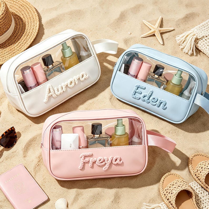 Puede incluir: Tres neceseres rectangulares en blanco, azul claro y rosa claro, cada uno con un panel frontal transparente y un asa lateral. Los bolsos est&aacute;n personalizados con los nombres Aurora, Eden y Freya en letras blancas en relieve y texturizadas.