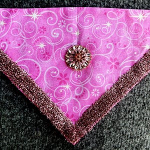 May include: A triangular pink fabric bandana featuring a white pattern of swirling lines, small flowers, and stars. The edges are trimmed with a textured, metallic rose-gold fringe, and a circular, faceted crystal brooch is centered on the fabric.