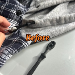 May include: A black plastic drawstring toggle replacement for a hooded garment. The toggle features a textured surface and a dual-hole design. One toggle is attached to a grey drawstring on a navy and white plaid hoodie, while another is loose.