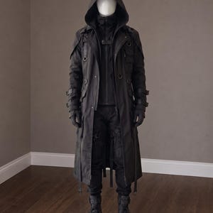 May include: A black hooded long coat featuring multiple straps, buckles, and D-ring hardware, worn over a black jacket and black trousers. The ensemble includes black gloves and black tactical-style boots, displayed on a white mannequin.