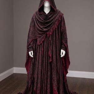 May include: A floor-length, dark maroon hooded cloak featuring a textured, distressed fabric with frayed edges. The garment includes a deep hood, long sleeves, and a layered, flowing design that drapes to the floor, revealing a lighter red inner lining.