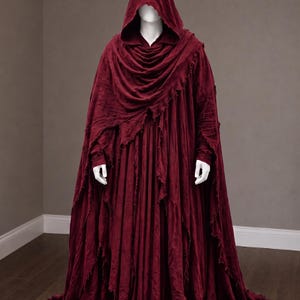 May include: A floor-length, deep burgundy hooded cloak featuring a textured, distressed fabric with frayed edges. The garment drapes loosely over the shoulders and body, creating a layered appearance with a pointed hood and long, flowing sleeves.