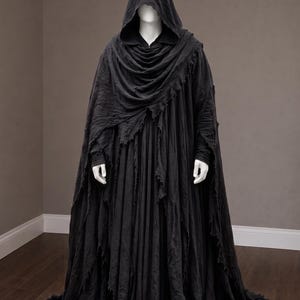 Pode incluir: Uma capa preta com capuz que chega ao chão, apresentando um design de tecido em camadas e desgastado com bordas desfiadas. A peça inclui um capuz profundo, mangas compridas e um corpete drapeado e plissado que se estende até o chão.