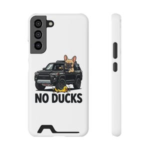 May include: A white smartphone case featuring a graphic of a tan French Bulldog driving a black SUV. A small yellow rubber duck lies on the ground in front of the vehicle. The text NO DUCKS is printed in bold black capital letters below the illustration.