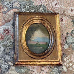 May include: A small rectangular gold-toned frame with an oval center opening containing a landscape painting. The painting depicts a green field, distant blue mountains, and a cloudy sky. A small metal hanging loop is attached to the top edge.