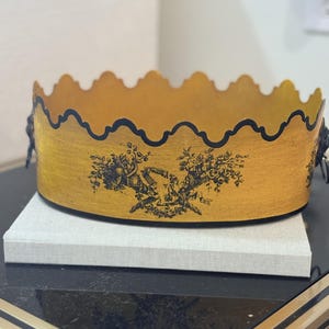 May include: An oval-shaped decorative tray with a metallic gold finish and a scalloped top edge outlined in black. The exterior features a detailed black floral transfer print. The tray includes small black metal side handles with ring pulls.