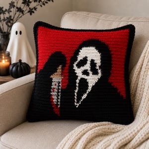 May include: A square crochet pillow featuring a black and white ghost-like figure with a screaming expression on a vibrant red background. The figure holds a silver knife with red accents, all rendered in detailed yarn stitches with a black border.