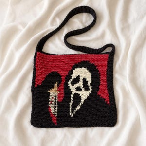 May include: A square crochet shoulder bag featuring a black and white ghost-like mask graphic on a red and black background. The design includes a small knife detail with red accents. The bag has a long, solid black crocheted shoulder strap.