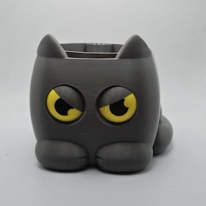 May include: A dark gray, cat-shaped container with a smooth, matte finish. The cat has two pointed ears, two large, round, yellow eyes with black pupils, and two front paws resting at the base. The top of the container is open.