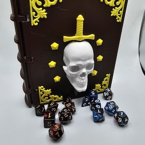 May include: A brown book-shaped storage box featuring yellow corner accents, a central white skull, and a yellow sword emblem. The box is surrounded by two sets of multi-sided polyhedral dice, one set in dark red and black, and the other in blue and black.