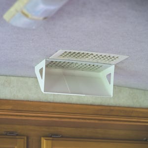 May include: A white plastic air vent deflector attached to a ceiling vent. The deflector features a rectangular grid pattern on the top section and a solid, angled front panel with triangular cutouts on the sides to redirect airflow.