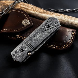 May include: A folding pocket knife with a grey and black Damascus steel handle featuring a distinct wavy pattern and a triangular geometric design. The knife is closed and rests on a brown leather sheath with a circular metal snap button.