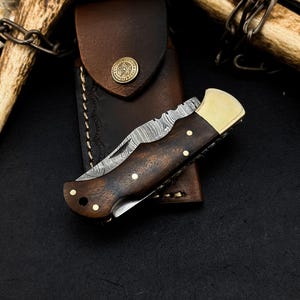 May include: A folding pocket knife with a Damascus steel blade, a dark brown wood handle with brass pins, and a brass bolster. The knife is positioned on a brown leather sheath featuring a circular brass button with a star emblem engraving.