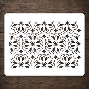 May include: A rectangular white stencil featuring a repeating pattern of black stylized floral motifs and small diamond shapes. The design consists of interconnected circular elements with curved petals arranged in a grid across the surface.