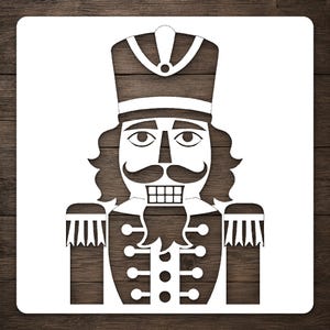 May include: A square stencil featuring a die-cut design of a nutcracker soldier. The nutcracker has a tall hat, a prominent mustache, a beard, and a uniform with epaulets and buttons. The design is white, creating a negative space image.