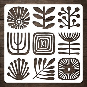 May include: A square white stencil featuring a grid of nine distinct cut-out shapes. The designs include various botanical-inspired patterns, such as stylized leaves, flowers with radiating petals, circular berry-like clusters, and concentric squares.