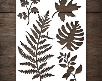 May include: A white rectangular stencil featuring five distinct botanical cutouts. The design includes a large central fern, a maple leaf, a monstera leaf, and two smaller leafy sprigs with rounded and pointed foliage shapes throughout.